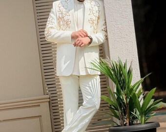 May include: Ivory suit with gold floral embroidery on the jacket. The suit includes a matching jacket, trousers, and a high-collared shirt. The person is wearing black loafers and sunglasses. The suit is a formal wear ensemble.