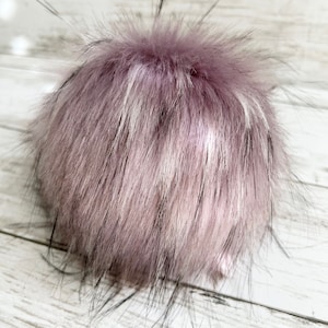 May include: A fluffy, round pompom in shades of lavender, white, and black. The faux fur has long, soft strands, creating a textured appearance. The pompom is likely for a hat or other craft project.