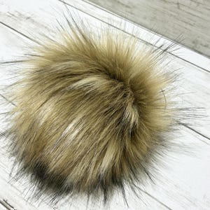 May include: A faux fur pom pom in shades of brown and black. The fluffy ball has long, textured fibers and a round shape. The pom pom is likely for hats, scarves, or other craft projects.