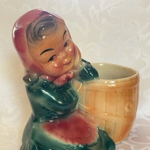 May include: A ceramic planter shaped like a girl wearing a red and green dress and a red bonnet. The planter has a brown and tan barrel-shaped pot attached to the front.