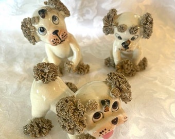 May include: Three white ceramic poodle figurines with brown yarn hair. The poodles are in various poses, with one looking directly at the camera.