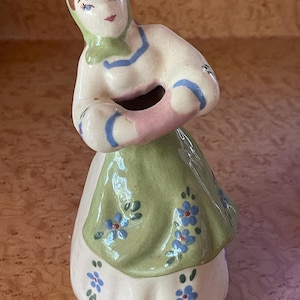 May include: A ceramic figurine of a woman in a green dress with blue flowers, a pink shawl, and a blue and white head scarf. The figurine has a hole in the center of the shawl, suggesting it may be a planter.