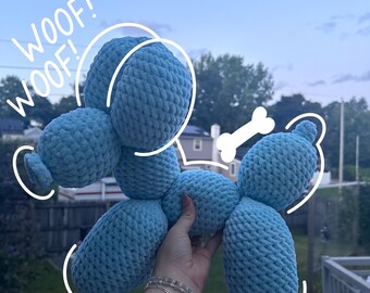 May include: A light blue crocheted dog toy in a balloon animal style. The toy has a bone-shaped detail and the words "WOOF! WOOF!" are written in white. The dog is held up by a person.
