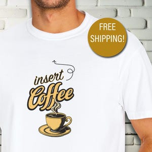 May include: White t-shirt featuring a coffee-themed graphic. The design includes the words "insert Coffee" above a steaming coffee cup and saucer. The graphic is in shades of yellow and brown.