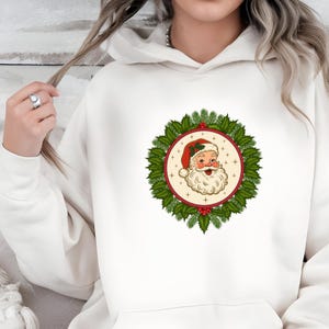 May include: White hooded sweatshirt featuring a circular graphic of Santa Claus. The Santa illustration is surrounded by a wreath of green leaves and red berries. Santa is wearing a red hat and has a white beard.