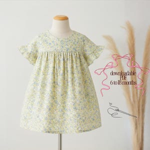 Puede incluir: Vestido de bebé amarillo con estampado floral de pequeñas flores azules. El vestido tiene mangas cortas con volantes y un corpiño fruncido. El texto "downloadable PDF 6 to 18 months" es visible en la imagen.