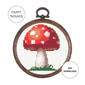 May include: A cross-stitch mushroom design in a wooden embroidery hoop. The mushroom has a red cap with white spots and a white stem, set against a white background. The hoop is brown, and the text "CRAFT BAZAAR" and "PDF DOWNLOAD" are visible.