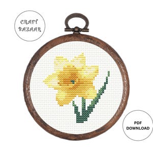 May include: A cross-stitch pattern of a yellow daffodil flower with green leaves in a wooden embroidery hoop. The pattern is available as a PDF download.