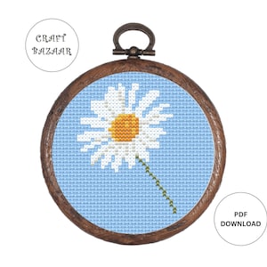 May include: A cross-stitch pattern of a white daisy with a yellow center on a light blue background. The pattern is framed in a brown wooden hoop.