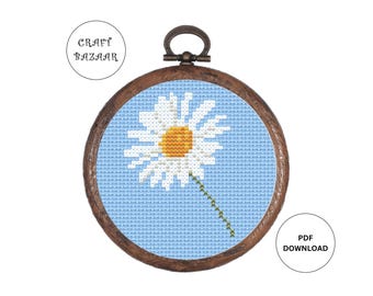 May include: A cross-stitch pattern of a white daisy with a yellow center on a light blue background. The pattern is framed in a brown wooden hoop.