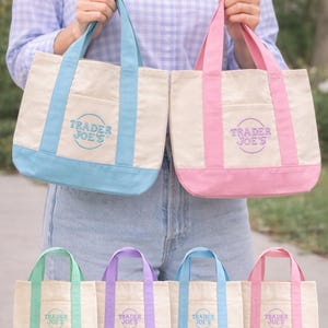 Könnte beinhalten: Eine Auswahl an Canvas-Tragetaschen in verschiedenen Farben, darunter Blau, Rosa, Grün und Lila. Jede Tasche hat einen kontrastierenden Boden und Griffe, mit dem Text "Trader Joe's" auf der Vorderseite gestickt. Die Taschen werden von einer Person gehalten.
