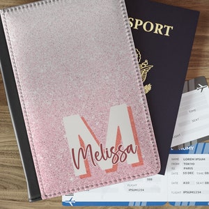 May include: A pink and white passport holder with a large letter "M" and the name "Melissa" printed on the front. The holder is sitting on top of a blue passport and a boarding pass.