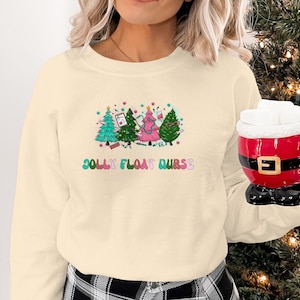 Peut inclure: Sweat-shirt crème avec un motif coloré d'arbres de Noël et le texte "JOLLY FLOAT NURSE". Les arbres sont décorés avec des fournitures médicales. Une personne tient une tasse de Père Noël rouge avec un dessus blanc.