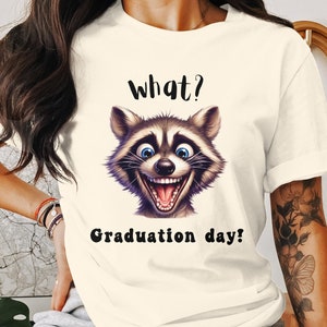 May include: White t-shirt with a cartoon raccoon face and the text "What? Graduation day!"