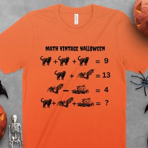 May include: Orange t-shirt with the text "MATH VINTAGE HALLOWEEN" and Halloween-themed math equations. The equations feature black cat and bat illustrations. The shirt is decorated with Halloween-themed decorations.