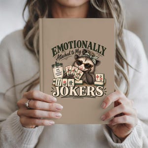 May include: A tan hardcover journal featuring a graphic of an opossum wearing a green headband and sunglasses, holding three joker playing cards. The text reads "EMOTIONALLY Attached to My JOKERS" with a tumbler labeled "SAME TILES DIFFERENT TRAUMA".