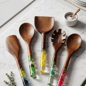 Puede incluir: Un juego de cinco utensilios de cocina de madera con mangos de resina colorida. Los mangos presentan diseños incrustados en azul, verde, amarillo y rojo. Los utensilios incluyen una cuchara, una espátula, una cuchara ranurada y dos cucharas más. Sobre superficie de mármol.