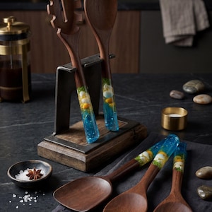 Puede incluir: Utensilios de cocina de madera con mangos de resina azul. El juego incluye una cuchara ranurada, una cuchara maciza, una espátula y dos cucharas más. Los mangos tienen resina azul transparente con elementos decorativos. Los utensilios se exhiben sobre una superficie oscura.