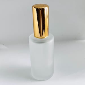 Könnte beinhalten: Eine parfümflasche aus mattiertem Glas mit goldfarbenem Deckel. Die Flasche hat eine zylindrische Form und einen abgerundeten Boden. Der Deckel ist goldfarben und rechteckig. Die Flasche ist leer.