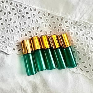 May include: Six green glass roller bottles with gold-colored caps are arranged diagonally on a white, eyelet-patterned fabric. The bottles are cylindrical and appear to be empty. The gold caps provide a contrast to the green glass.