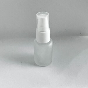 May include: A small, frosted glass spray bottle with a white pump and clear cap. The bottle is cylindrical and appears empty, suitable for essential oils or perfumes. The background is a plain, light gray.