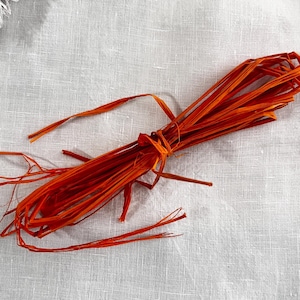 May include: A bundle of orange raffia tied with twine, set against a white linen backdrop. The raffia strands are long and thin, with a slightly frayed texture. Suitable for crafting and decorative use.