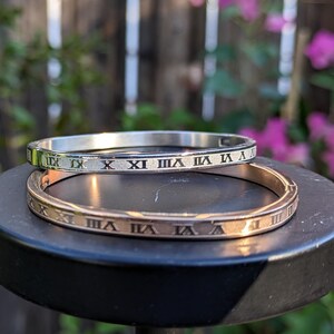 May include: Two bangle bracelets, one silver and one rose gold, with Roman numeral engravings. The bracelets are 7 inches or 17.78 centimeters in diameter.