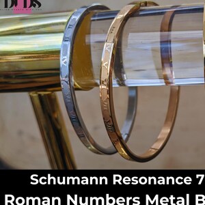 May include: Three metal bracelets with Roman numeral engravings. The bracelets are displayed on a clear acrylic stand. The bracelets are gold, silver, and rose gold in color. The text "Schumann Resonance 7.83Hz Roman Numbers Metal Bracelets" is displayed below the bracelets.