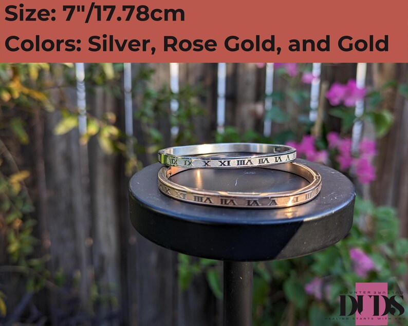 May include: Two bangle bracelets, one silver and one rose gold, with Roman numeral engravings. The bracelets are 7 inches or 17.78 centimeters in diameter.