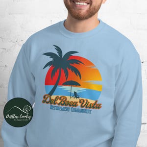 May include: A light blue sweatshirt with a graphic design of a palm tree on a beach with a sunset in the background. The text "Del Boca Vista Retirement Community" is printed below the graphic.