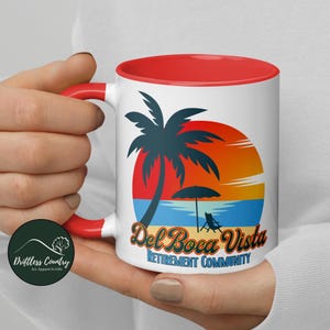 May include: White ceramic mug with a red rim. The mug features a colorful illustration of a palm tree on a beach with a sunset in the background. The text "Del Boca Vista Retirement Community" is printed on the mug.