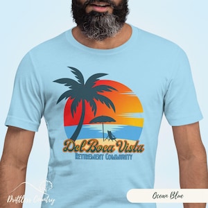 May include: A light blue t-shirt with a graphic of a palm tree on a beach with a sunset in the background. The text "Del Boca Vista Retirement Community" is printed on the shirt.