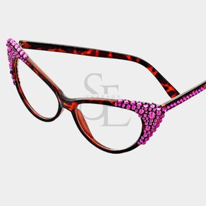May include: Cat-eye style eyeglasses with a brown and black tortoiseshell frame. The upper corners of the frames are embellished with pink rhinestones. The lenses are clear.
