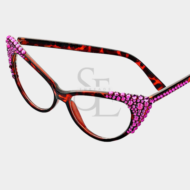 May include: Cat-eye style eyeglasses with a brown and black tortoiseshell frame. The upper corners of the frames are embellished with pink rhinestones. The lenses are clear.