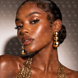 May include: A close-up shot of a person wearing gold earrings. The earrings are composed of three stacked spheres. The person is wearing a gold sequined top. The person has dark skin, dark hair, and is wearing makeup.
