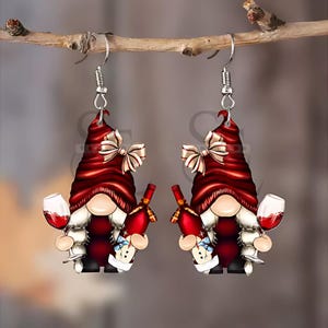 May include: A pair of dangle earrings featuring gnome figures with red hats and white beards. Each gnome holds a wine glass and a wine bottle. The earrings have silver-colored hooks and are suspended from a small branch.