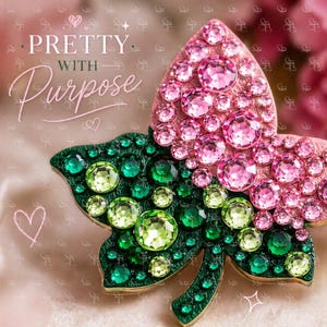 May include: A decorative leaf-shaped brooch with a pink and green gemstone design. The top half features pink gemstones, while the bottom half is green. The brooch has the words "Pretty with Purpose" in the upper left corner.