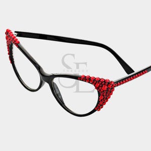 May include: Black cat-eye eyeglasses with red rhinestone accents. The frames are black, and the rhinestones are clustered along the outer edges of the frames. The lenses are clear.