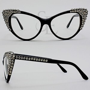 May include: Black cat-eye eyeglasses with clear lenses. The frames are embellished with numerous clear rhinestones along the upper outer edges. The glasses have a sleek, modern design.