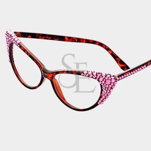 May include: Cat-eye style eyeglasses with a brown tortoiseshell frame. The outer edges of the frames are embellished with pink rhinestones. The glasses have clear lenses and are set against a white background.