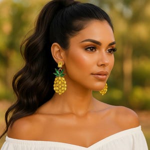 May include: Gold-colored pineapple earrings with green leaves and a square yellow gem at the top. The earrings are a tropical-themed accessory. The woman in the image is wearing a white off-the-shoulder top.