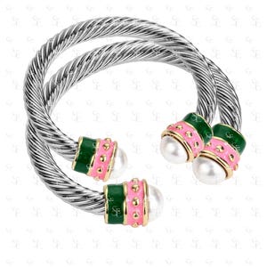 May include: Two silver-tone, twisted-cable bracelets. Each bracelet features green and pink accents with gold-colored details and a faux pearl. The bracelets are a stylish accessory.
