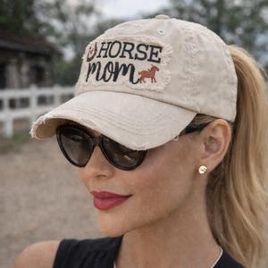 May include: A distressed beige baseball cap with the words "HORSE MOM" embroidered on a patch. The patch includes a red horse graphic. The cap has a pre-curved bill and a ponytail opening.