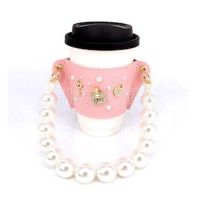 May include: Pink coffee cup sleeve with a pearl chain strap and gold accents. The sleeve is decorated with small pearls and gold charms.