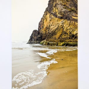 May include: A landscape print featuring a coastal scene. The image shows a sandy beach with foamy waves and a large, textured rock formation. The rock is a mix of yellow, brown, and black hues, contrasting with the overcast sky.