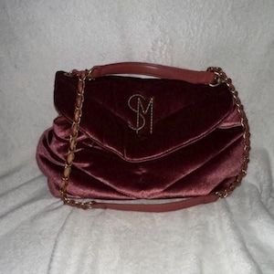May include: A deep burgundy velvet handbag featuring a quilted texture and a gold-toned chain strap with a matching velvet shoulder pad. The front flap displays a prominent gold-colored monogram logo consisting of the letters S and M intertwined.