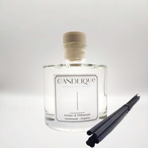 May include: A clear glass bottle with a wooden cap and a label that reads "CANDLIQUE ARTISANAL DECO Amber & Olibanum Handmade - Organic". The bottle is filled with a clear liquid and is sitting on a white surface. There are black diffuser sticks next to the bottle.