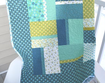 May include: A patchwork quilt in shades of teal, blue, and yellow, draped over a white chair. The quilt features various floral and geometric patterns, with a patterned border. The quilt is made of soft fabric.