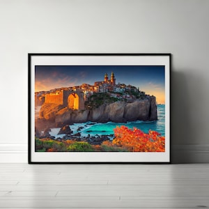 Puede incluir: Una fotografía de paisaje enmarcada que muestra un pueblo costero sobre un acantilado rocoso al atardecer. La escena presenta edificios de piedra, un arco prominente, agua turquesa y follaje naranja vibrante en primer plano.