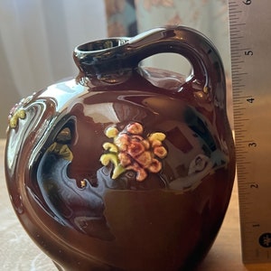 May include: A brown ceramic pitcher with a handle and floral accents. The pitcher has a glossy finish and a dark brown handle. The floral accents are in shades of yellow, pink, and green. The pitcher is approximately 6 inches tall.